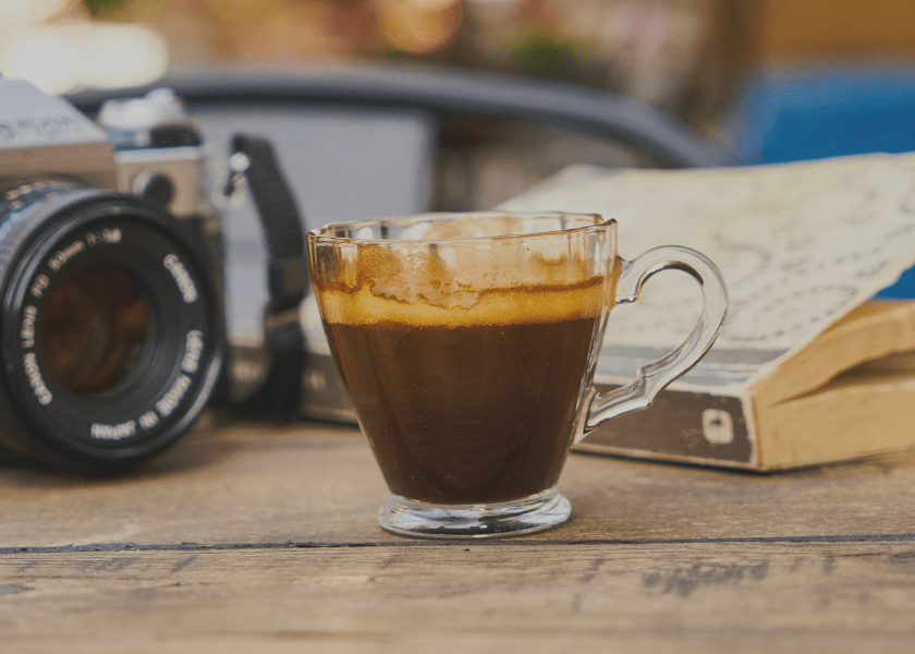 Ein Kaffee steht auf einem Tisch, im Hintergrund ein Buch und eine Kamera