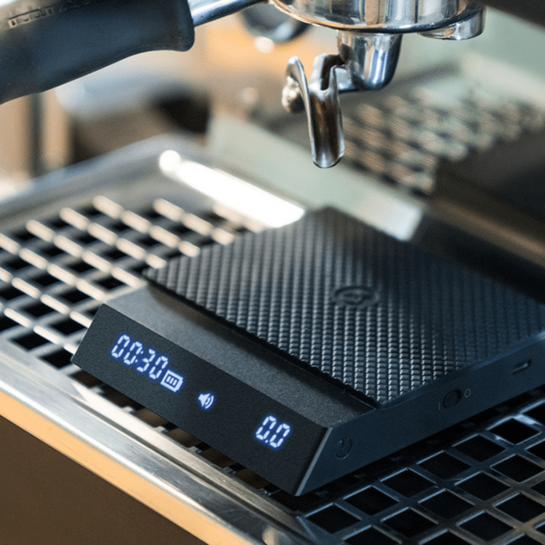 Die Timemore Black Mirror Nano Espresso Waage steht unter einer Siebträgermaschine mit dem Hitzschutzpad.
