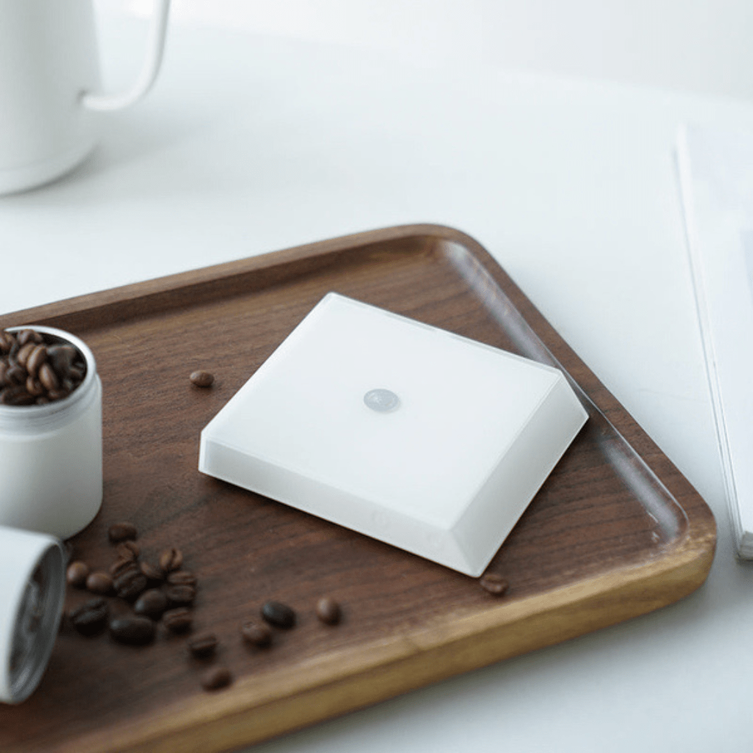 Die Timemore White Mirror Nano Espresso Waage steht auf einem Tablett, neben ihr sind Kaffeebohnen.