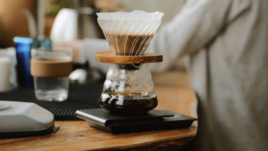 Ein Kaffeezubereiter steht auf einer Kaffeewaage. Die Kaffeewaage ist schwarz und steht wiederum auf einer Arbeitsplatte. Im Hintergrund ist eine Frau an einem Wasserhahn über die Spüle gebeugt. 