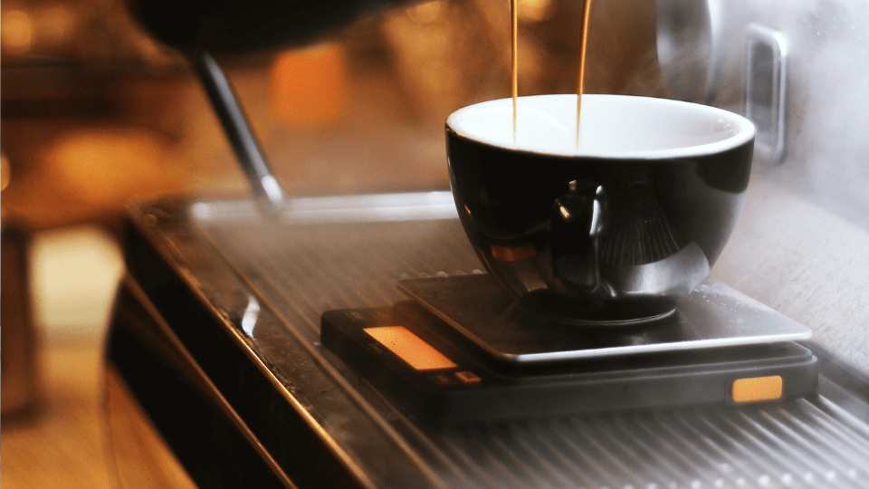 Eine Kaffeewaage steht unter einer Siebträgermaschine. Die Siebträgermaschine ist gerade in Betrieb und brüht Kaffee. Der Kaffee läuft in eine schwarze Tasse, die auf der Kaffeewaage positioniert ist. 